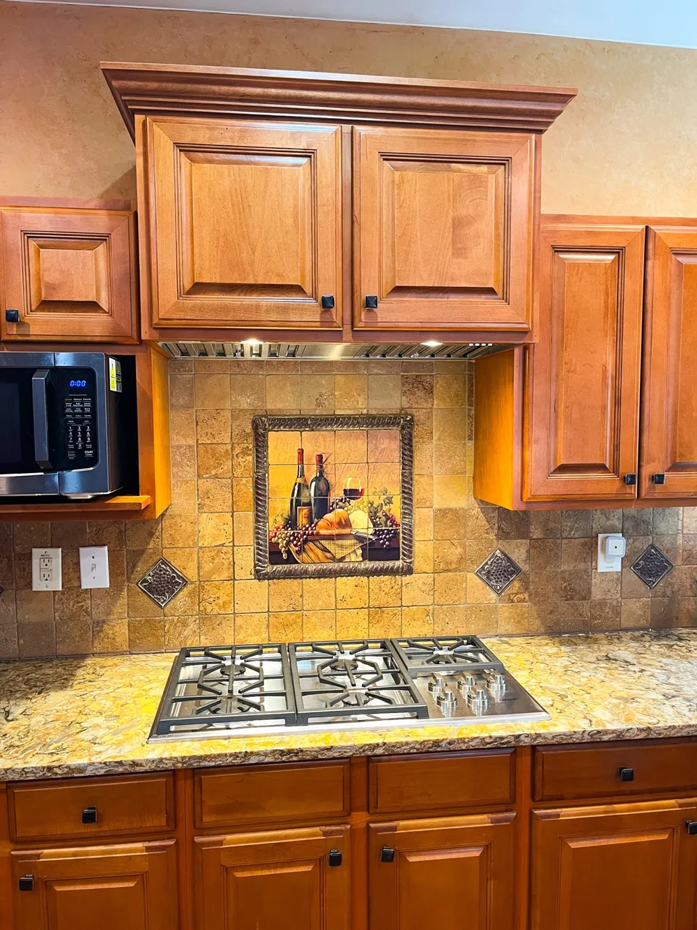 Wood cabinets, a gas cooktop and a stone tile backsplash with a tile mural above it